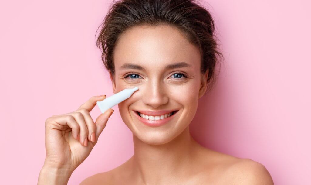 Beautiful woman using cream around the eyes. Photo of woman with perfect makeup on pink background. Beauty and skin care concept