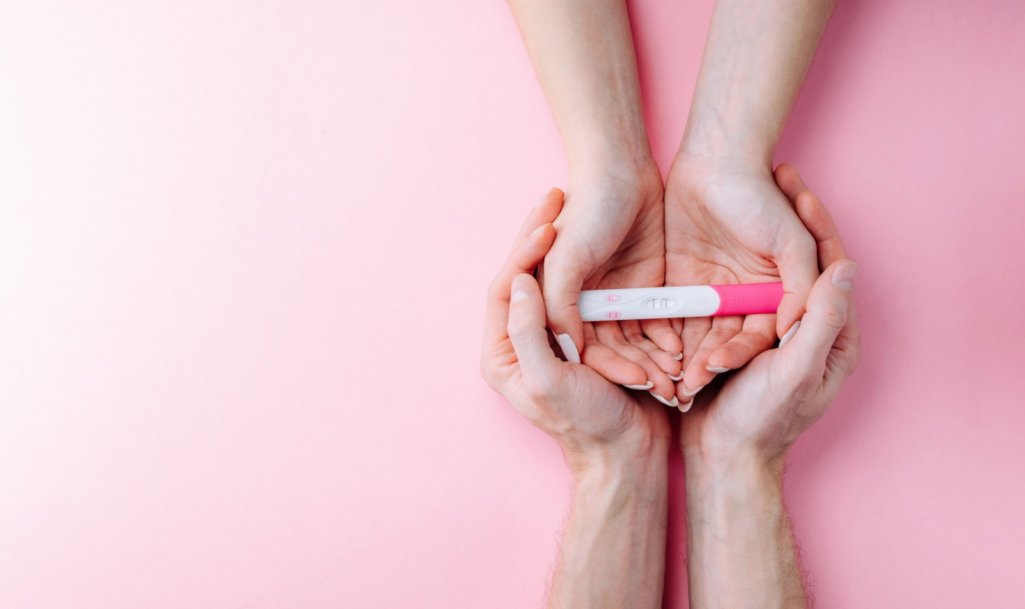 Female hand hold positive pregnant test with silk ribbon on pink banner background.
