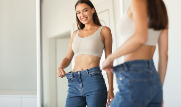 Slimming Concept. Excited Slim Woman Wearing Jeans After Great Weight Loss Comparing Size Posing Standing Near Mirror At Home. Selective Focus, Cropped Shot